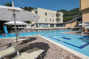 A swimming pool with lounge chairs and umbrellas, a building with balconies, and a mountainous backdrop.