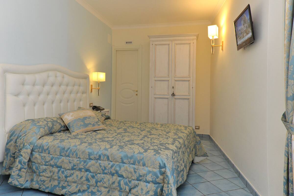 A bedroom with a tufted headboard, a floral bedspread, a bedside table with a lamp, and a door with a light switch.
