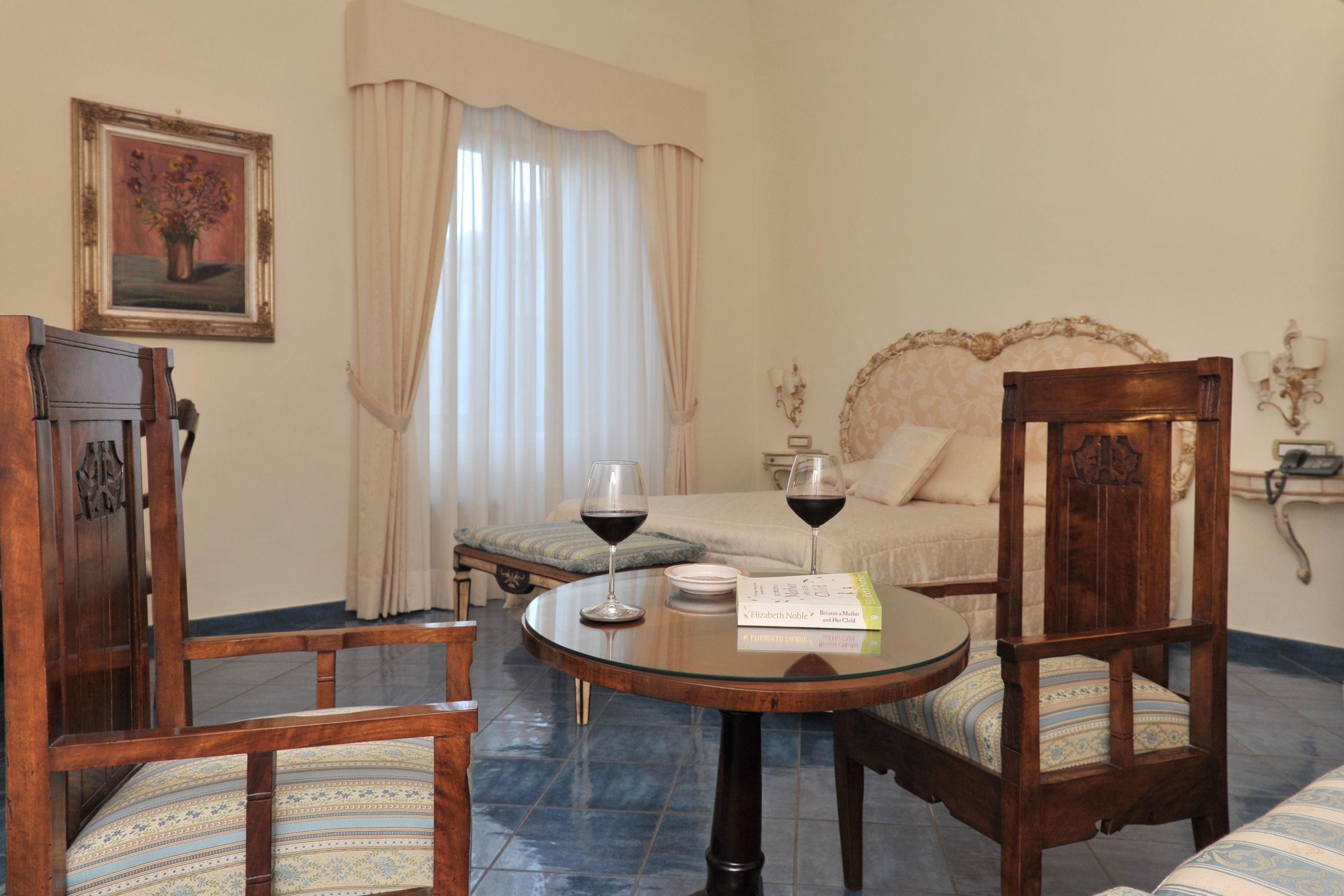 A room with a bed, a wooden chair, a round table with two wine glasses, a framed picture on the wall, and a bedside table with a lamp.