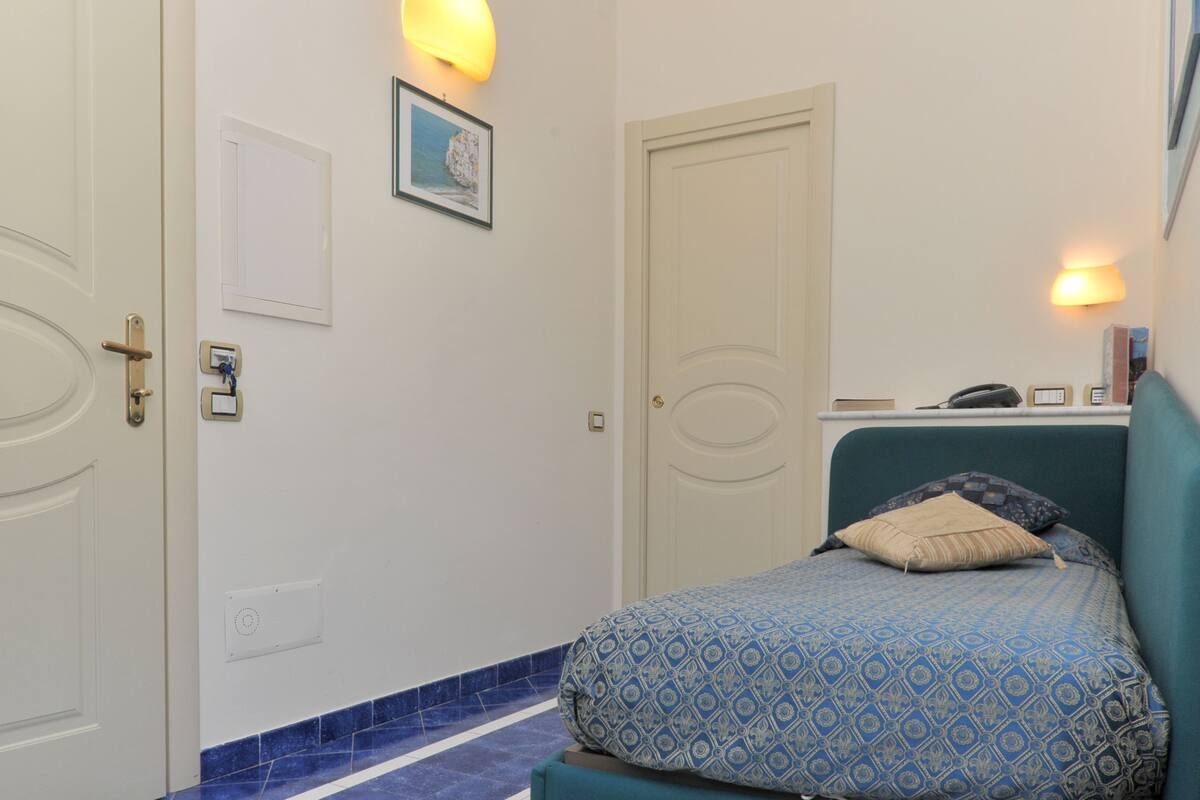 A bedroom with a bed, a door, a wall-mounted light, and a small shelf with items.