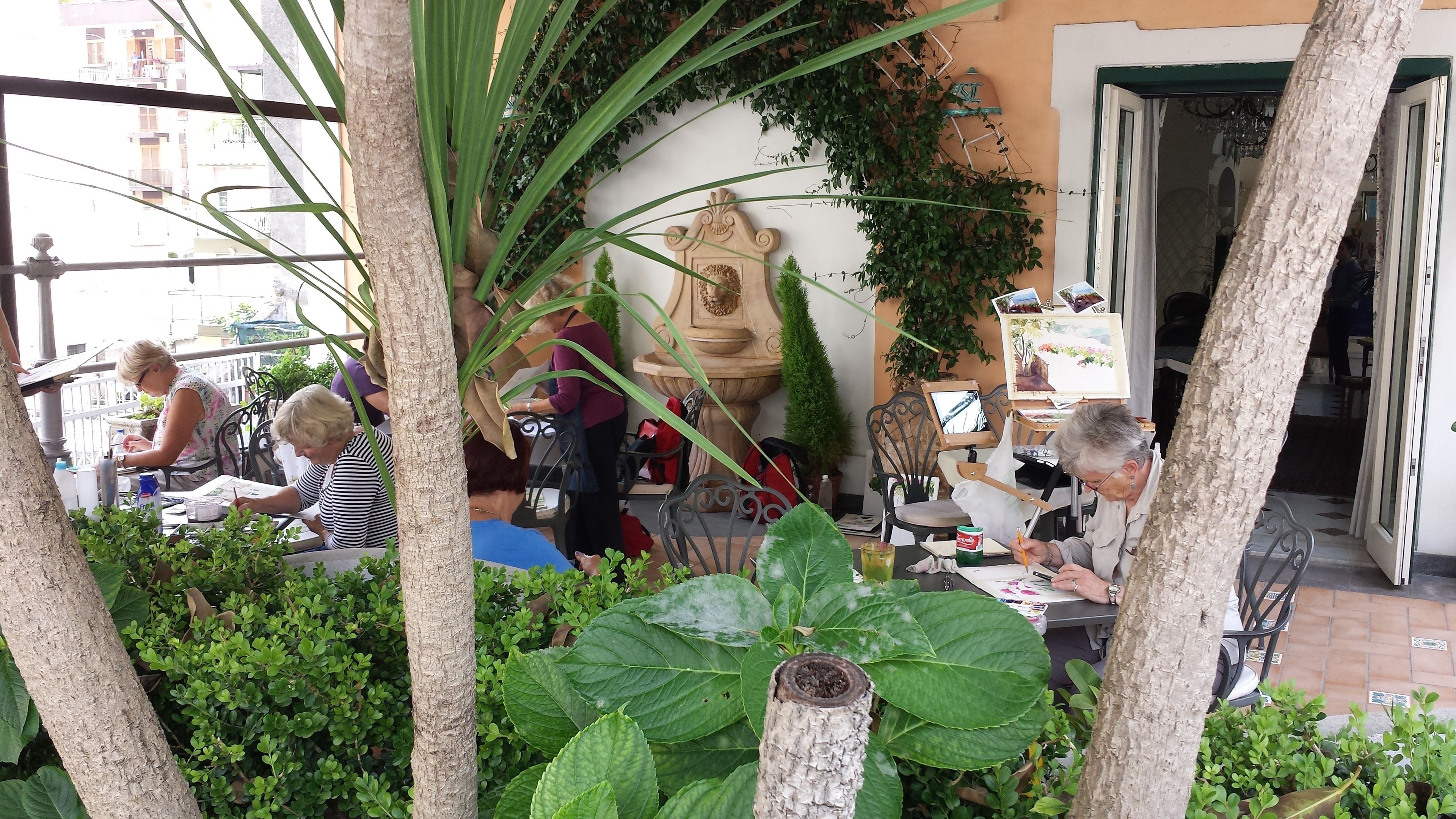 An outdoor café with tables and chairs, surrounded by greenery and a decorative stone fountain.