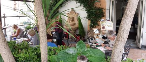 An outdoor café with tables and chairs, surrounded by greenery and a decorative stone fountain.