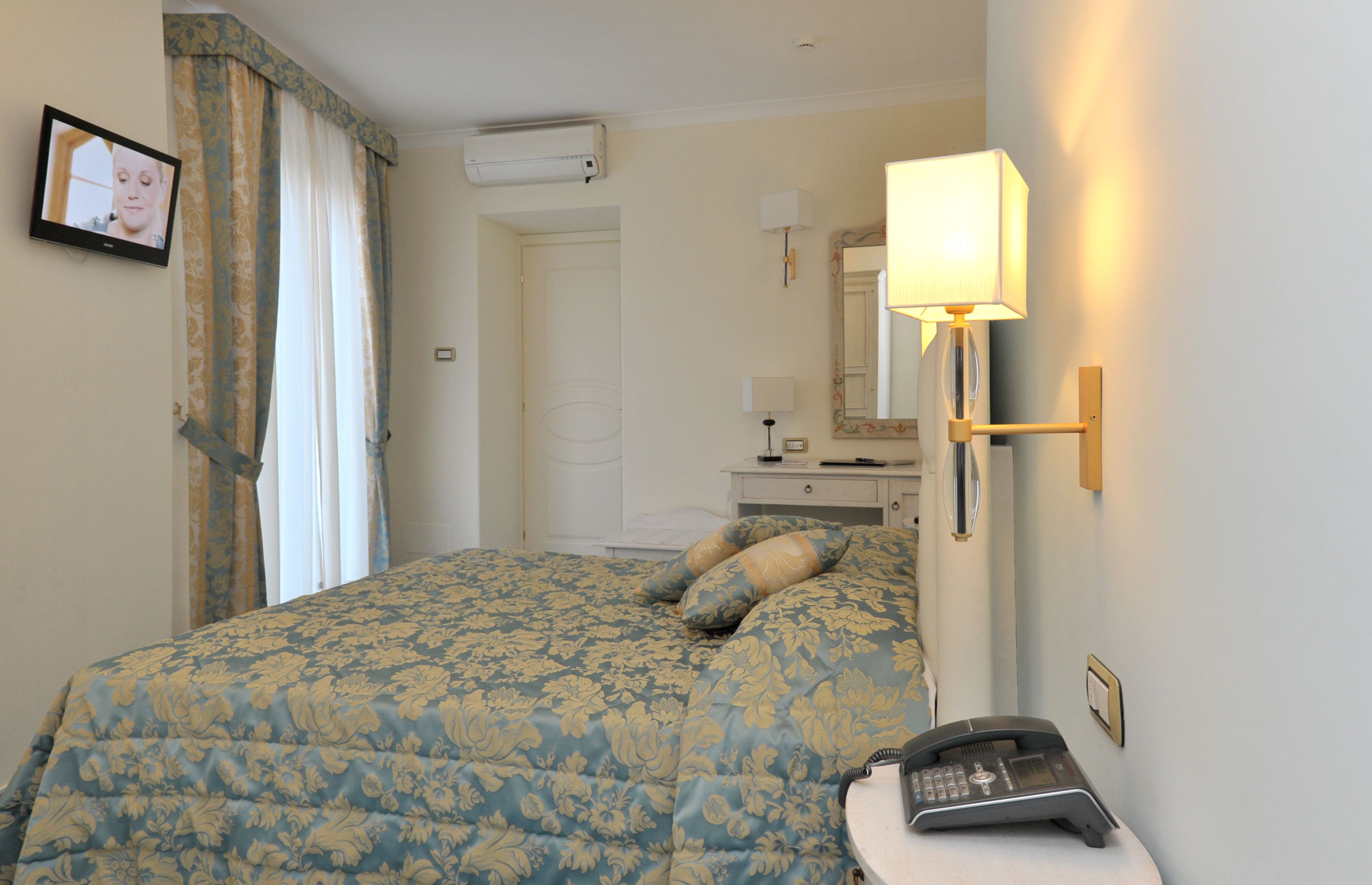 Habitación de hotel con cama, mesita de noche con teléfono, lámpara, televisor y espejo.