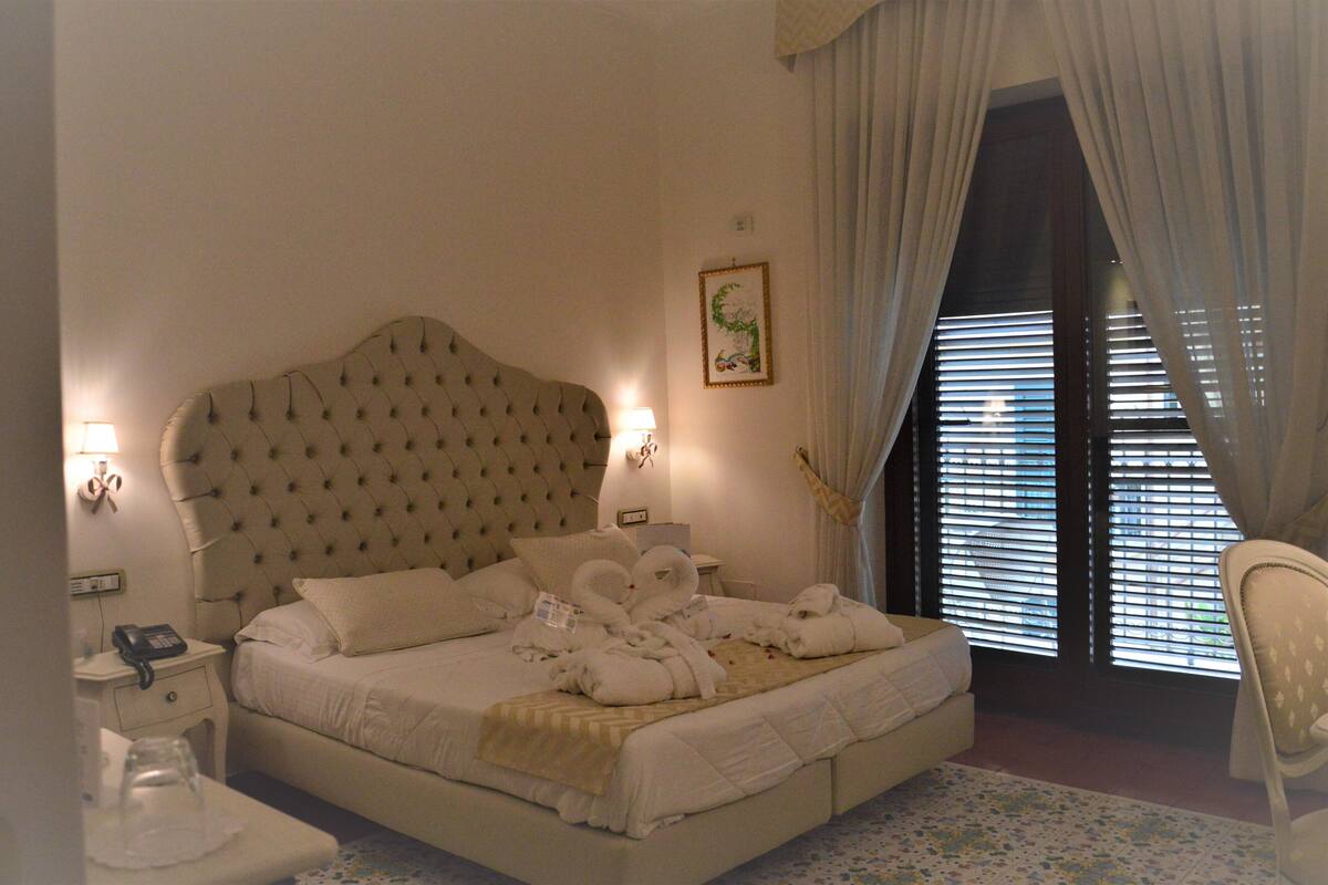 A hotel room with a large bed, a bedside table with a lamp, a chair, and a window with blinds.