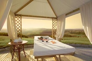 Un espace de détente extérieur aménagé sous une tente, comprenant une table de massage, des serviettes et une table avec une bouteille et des fruits.