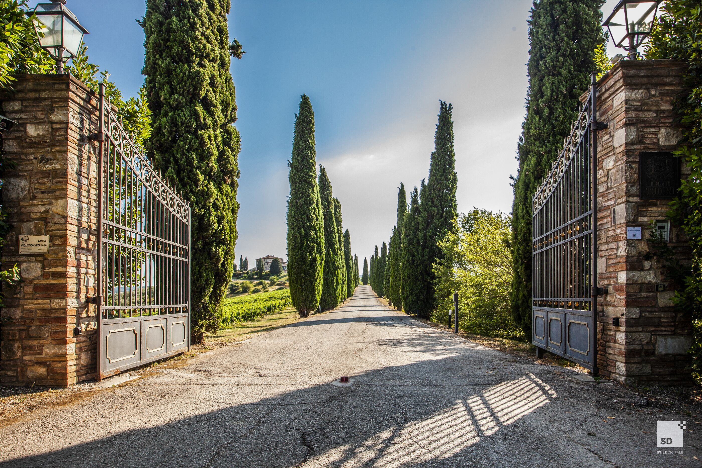Un chemin de gravier bordé de cyprès imposants et de portails en métal, qui mène à un bâtiment en pierre.
