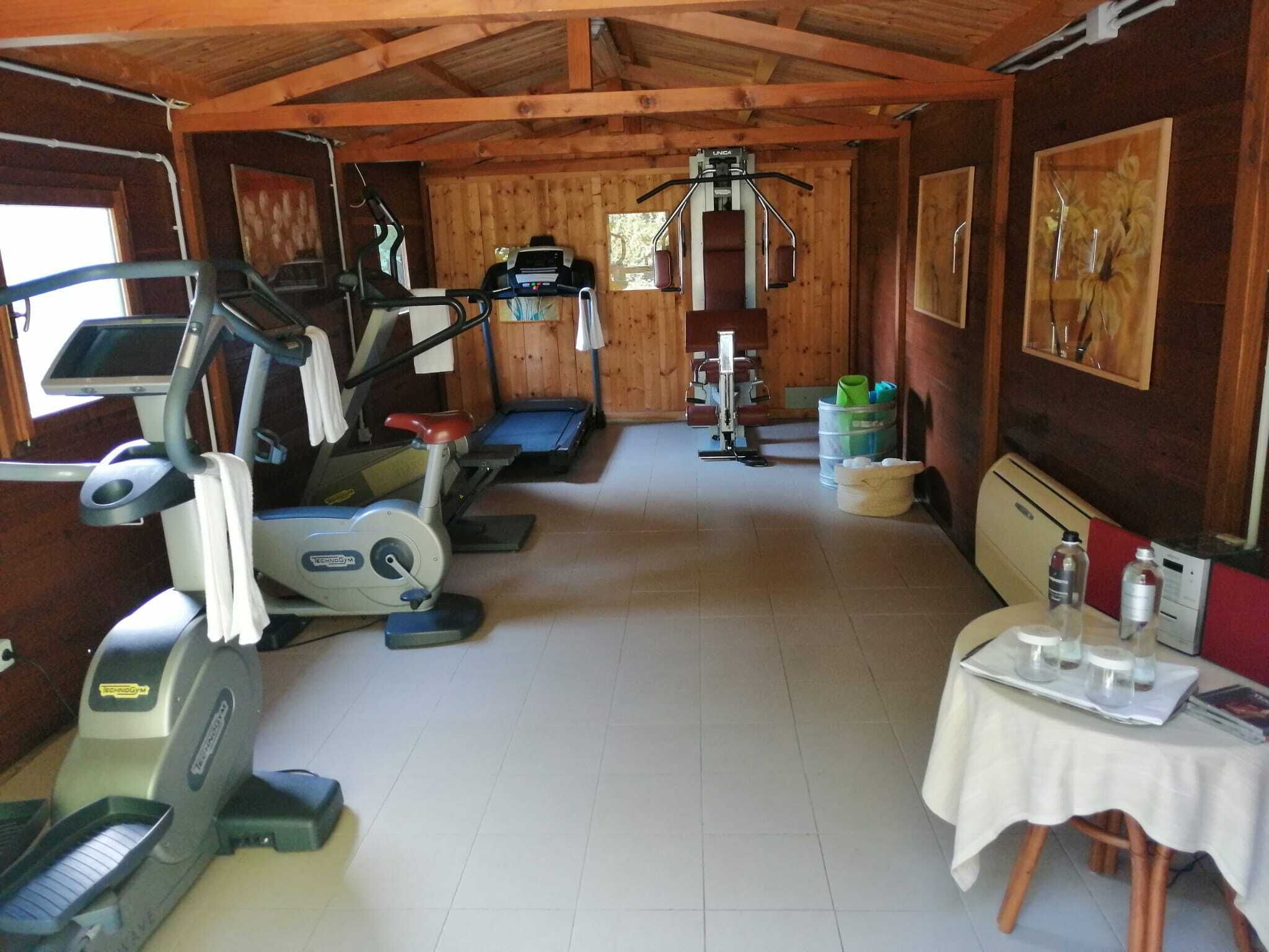 Il s'agit d'une salle de sport équipée de divers appareils, notamment des tapis de course, un vélo elliptique et un vélo d'appartement. On y trouve une table avec des bouteilles d'eau et une chaise. Les murs de la pièce sont en bois et le sol est carrelé.