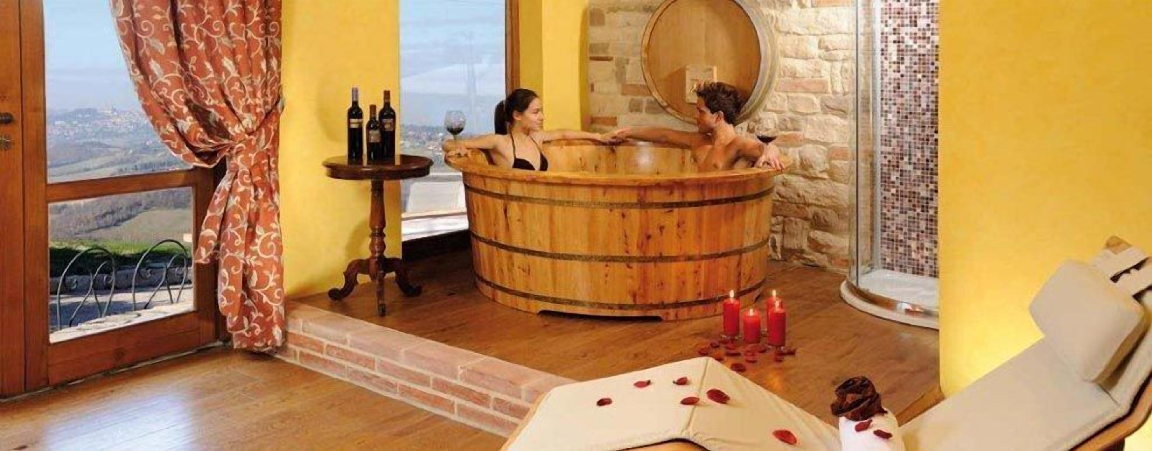 Una bañera de madera en un cuarto con vista a un paisaje urbano, una mesita con botellas y velas en el suelo.