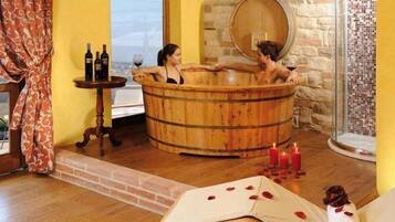 Una bañera de madera en un cuarto con vista a un paisaje urbano, una mesita con botellas y velas en el suelo.