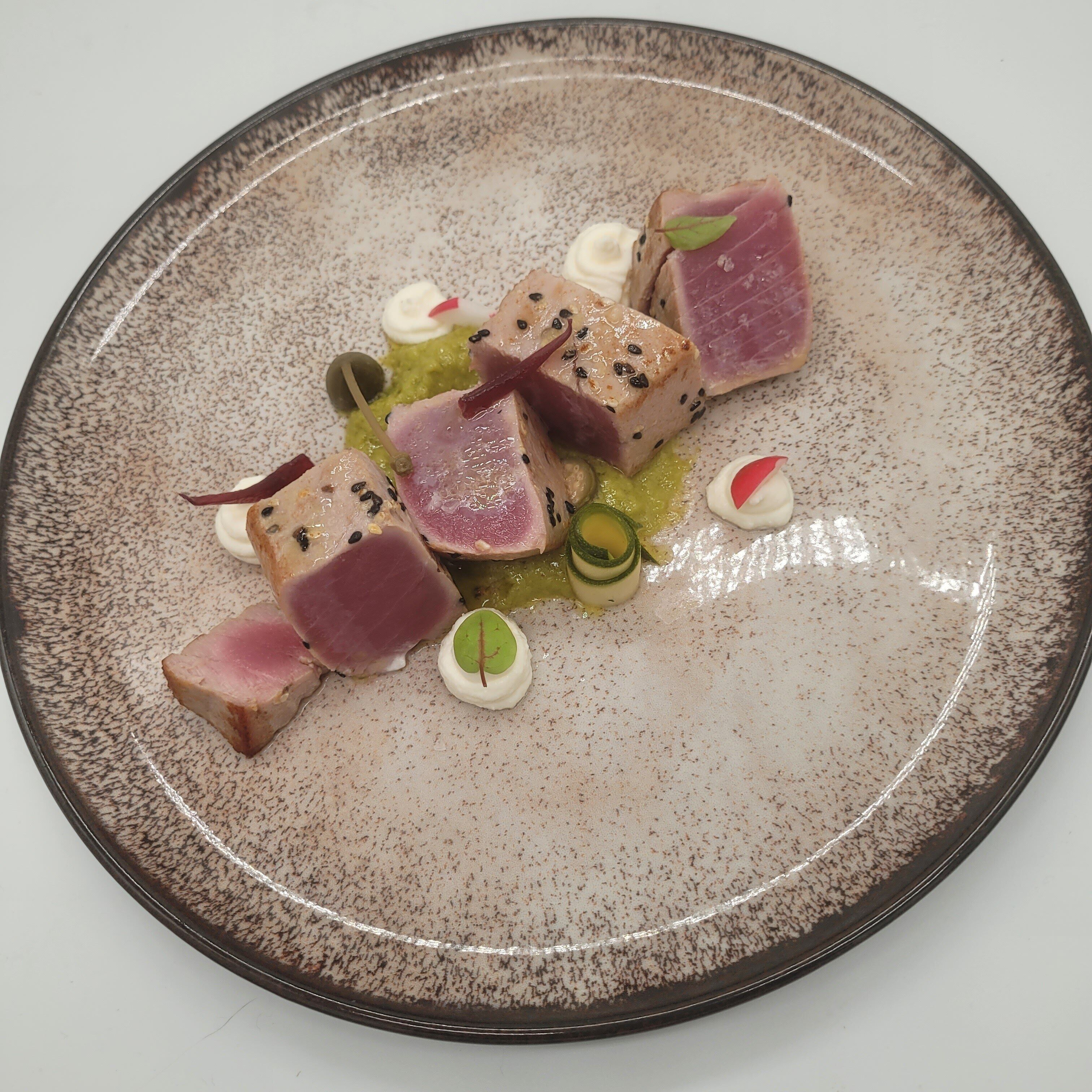 Un plato de cerámica redondo con un diseño moteado, que contiene varios trozos de carne rosada, hojas verdes y pequeños adornos blancos y rojos.