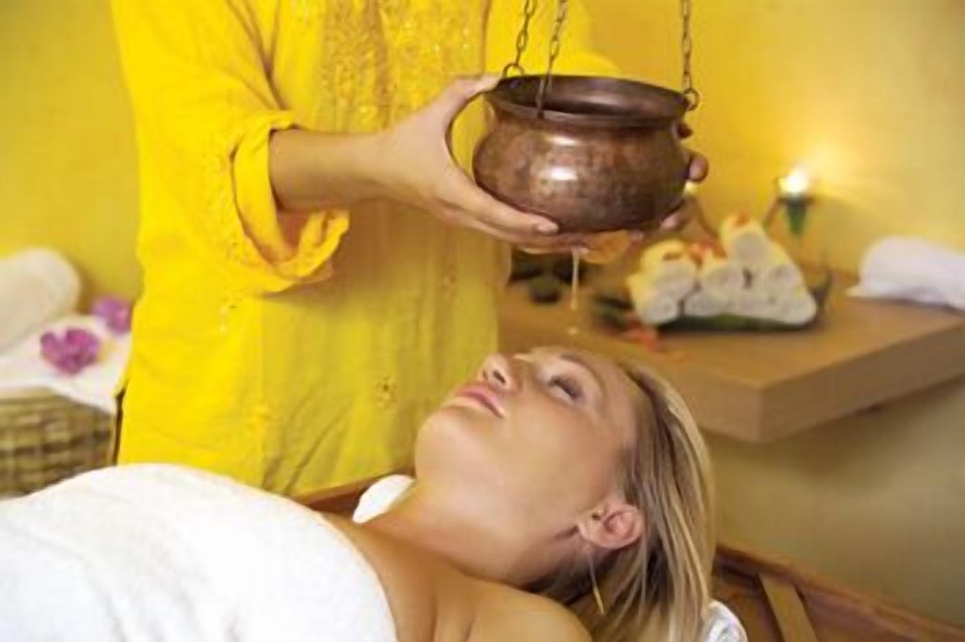 Uma pessoa com um robe amarelo segurando um pote, provavelmente para um tratamento de spa, com uma pessoa relaxada deitada em primeiro plano.