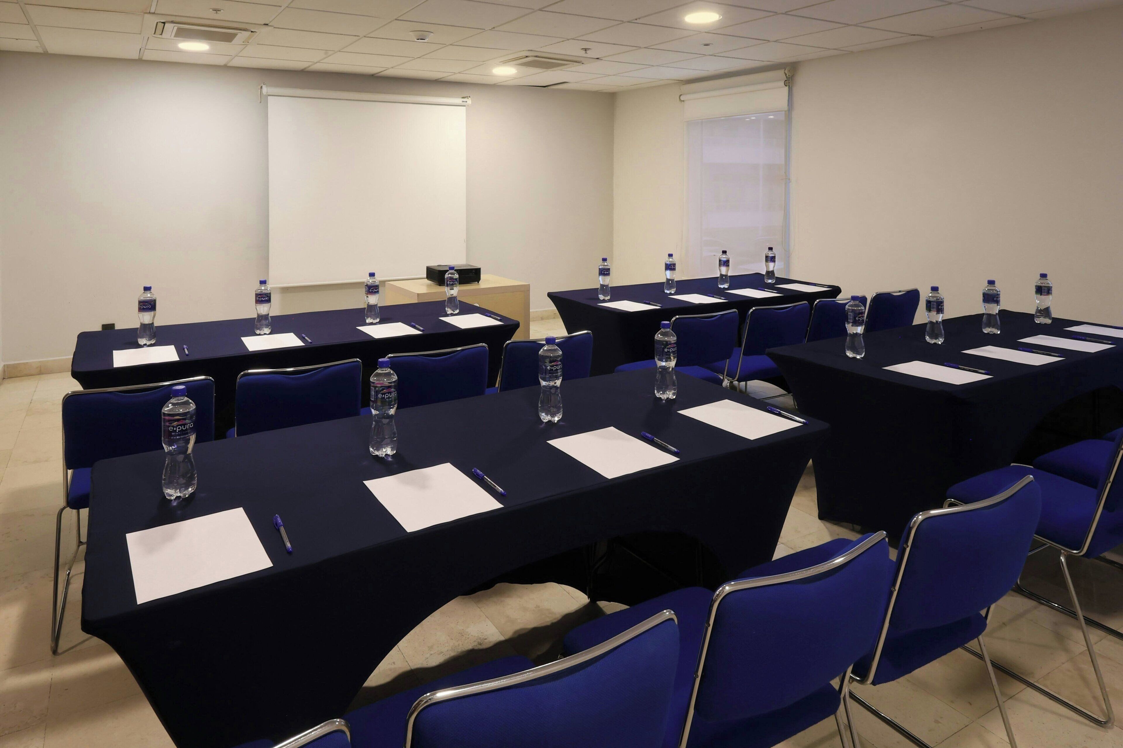 A conference room with multiple tables, chairs, water bottles, and a projector screen.