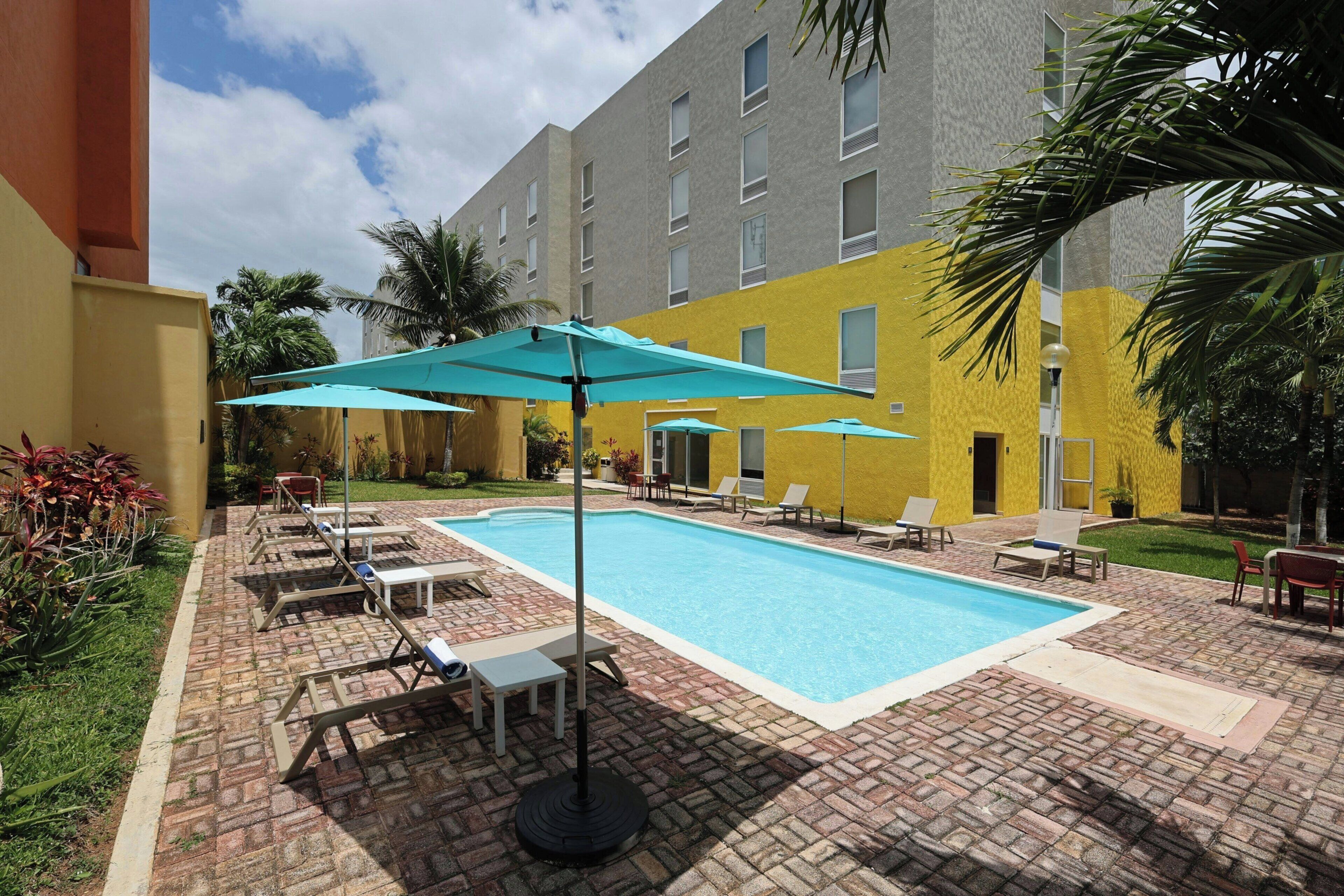A swimming pool with lounge chairs and umbrellas, surrounded by a brick patio and a yellow building.