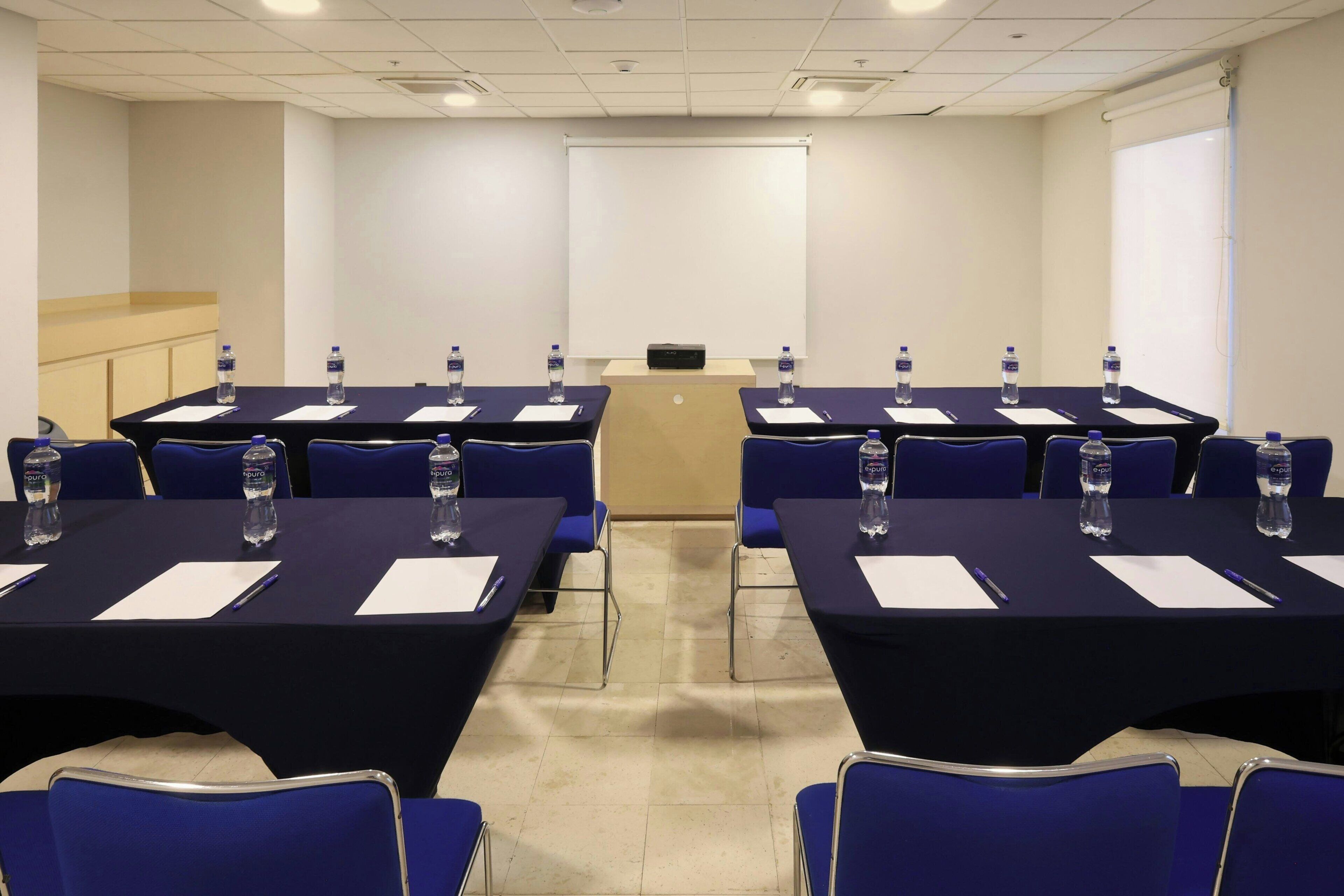 Una sala de conferencias con mesas, sillas, botellas de agua y una pantalla para proyector.