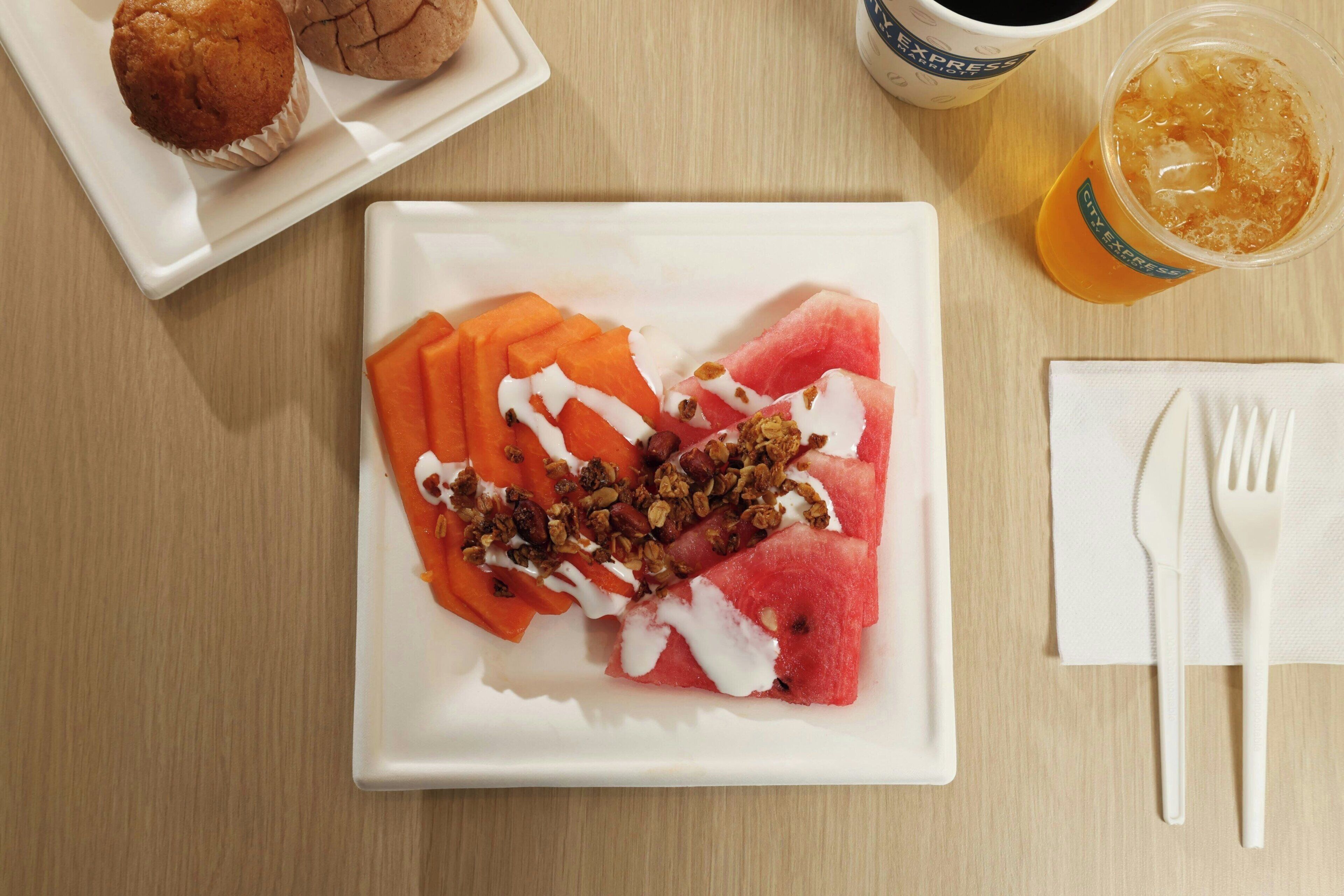 A plate of watermelon with yogurt and granola, a muffin, a cup of coffee, and a glass of iced tea on a wooden table.