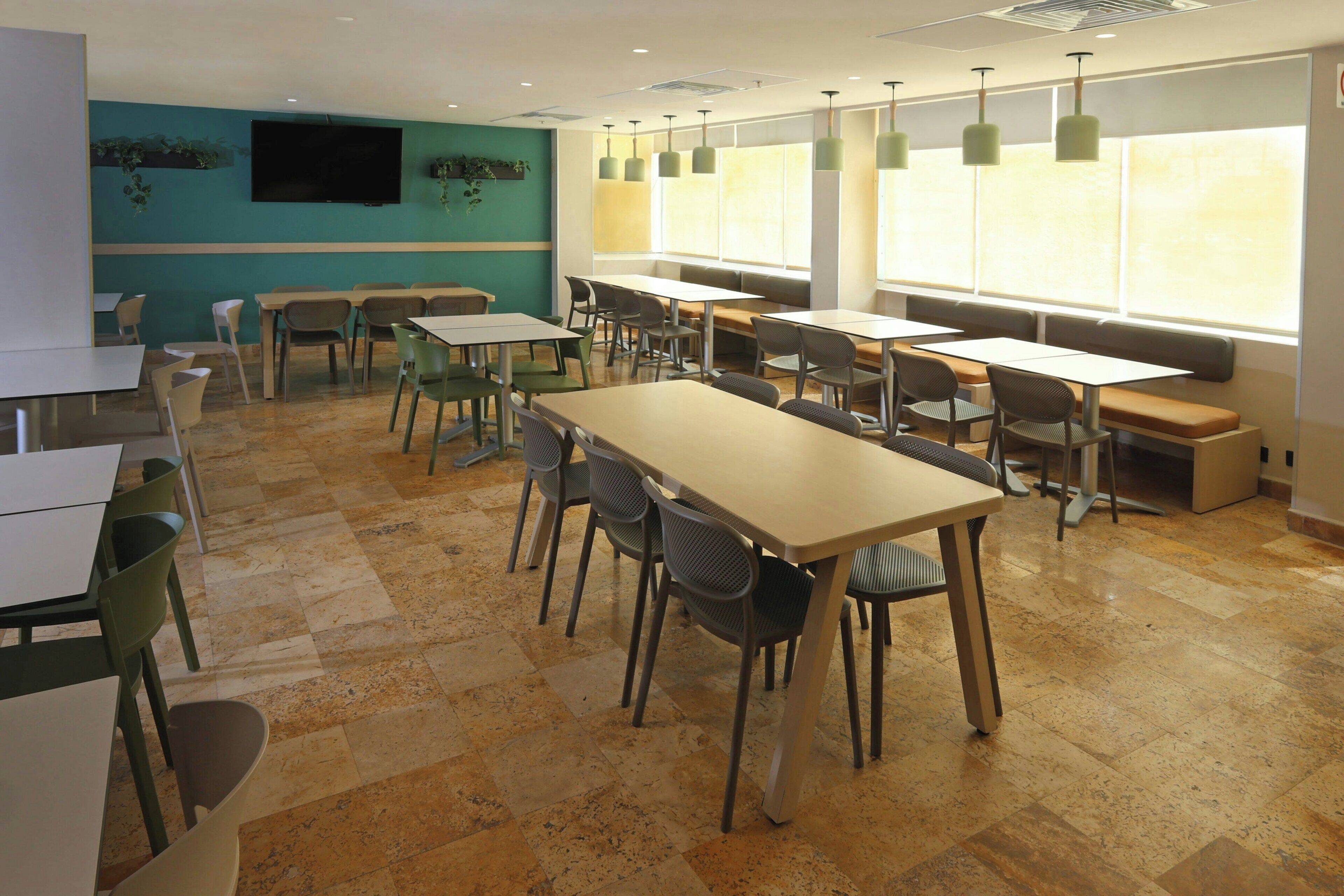 A dining area with multiple tables and chairs, a flat-screen TV mounted on the wall, and large windows allowing natural light.