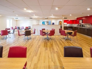 A modern dining area with red chairs and tables, a kitchen area with red cabinets, and a counter with a coffee machine. - Travelodge Manchester Sportcity (Manchester)