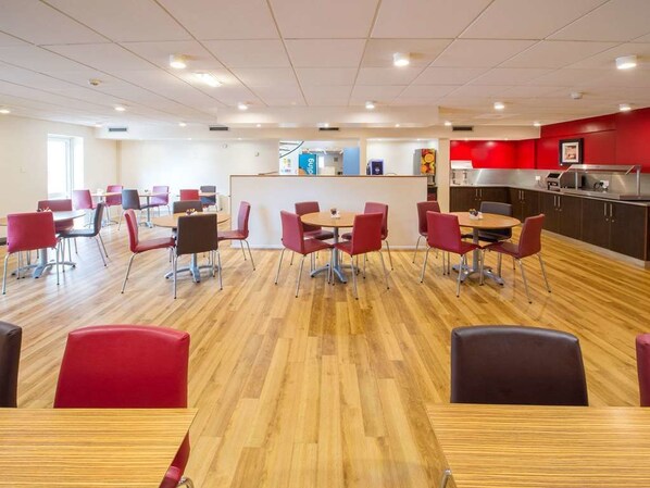 Un espace repas moderne avec des chaises rouges et noires, des tables en bois et un coin cuisine en arrière-plan.