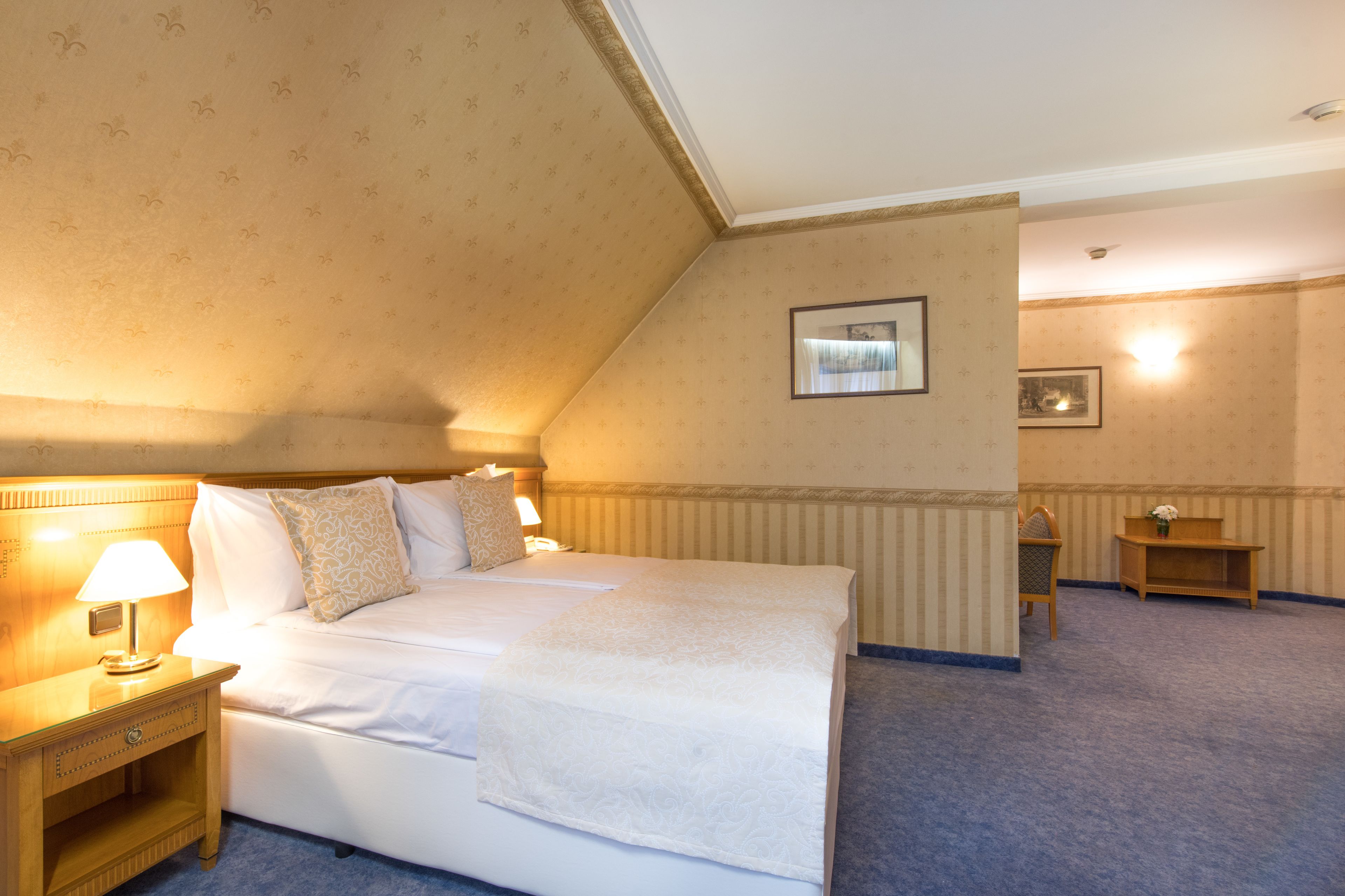 Una habitación de hotel con una cama grande, una mesita de noche con lámpara, un cuadro en la pared y una pequeña mesa de madera.