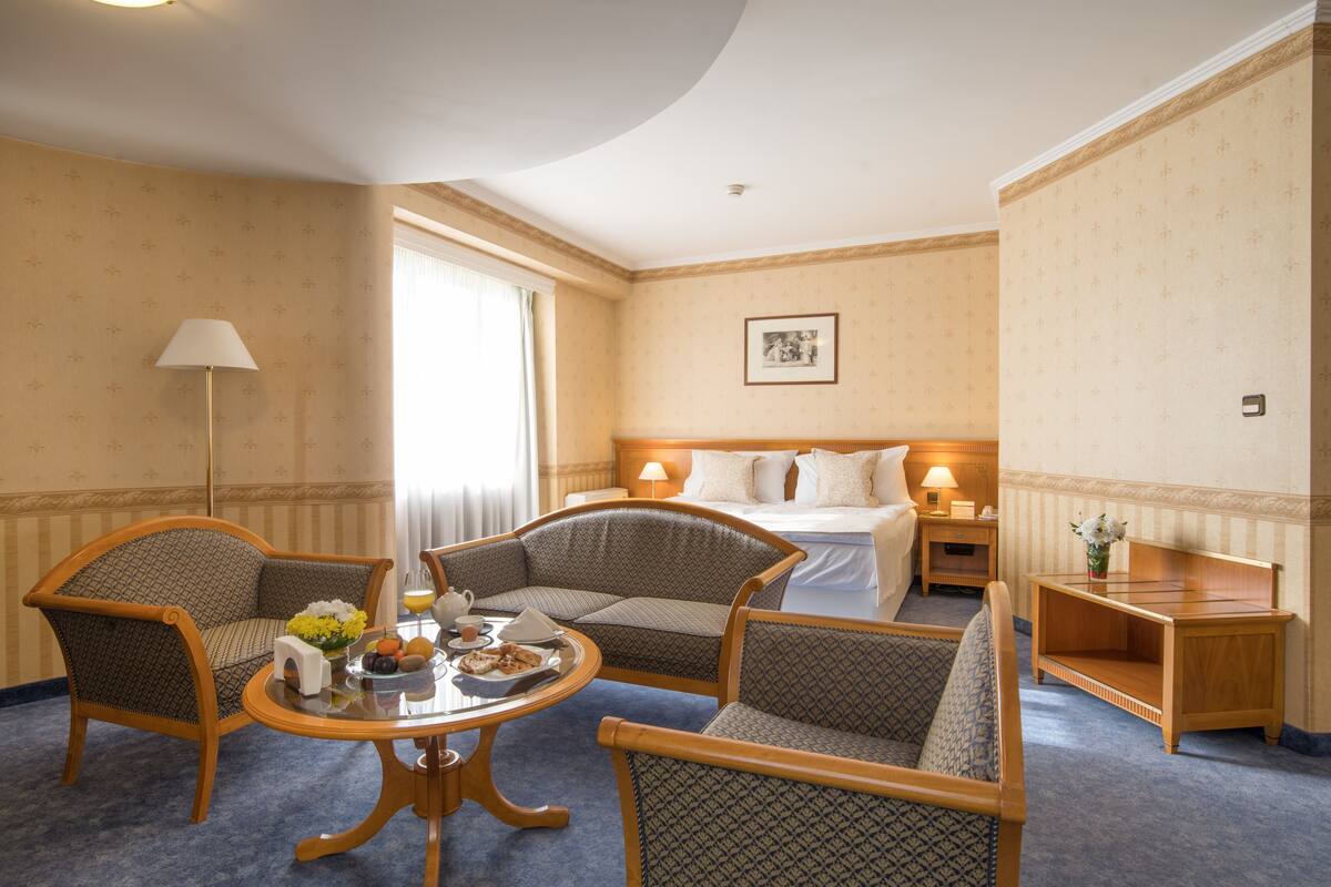A hotel room with a bed, two armchairs, a small table with a tray of fruit and a teapot, a nightstand with a lamp, and a picture on the wall.
