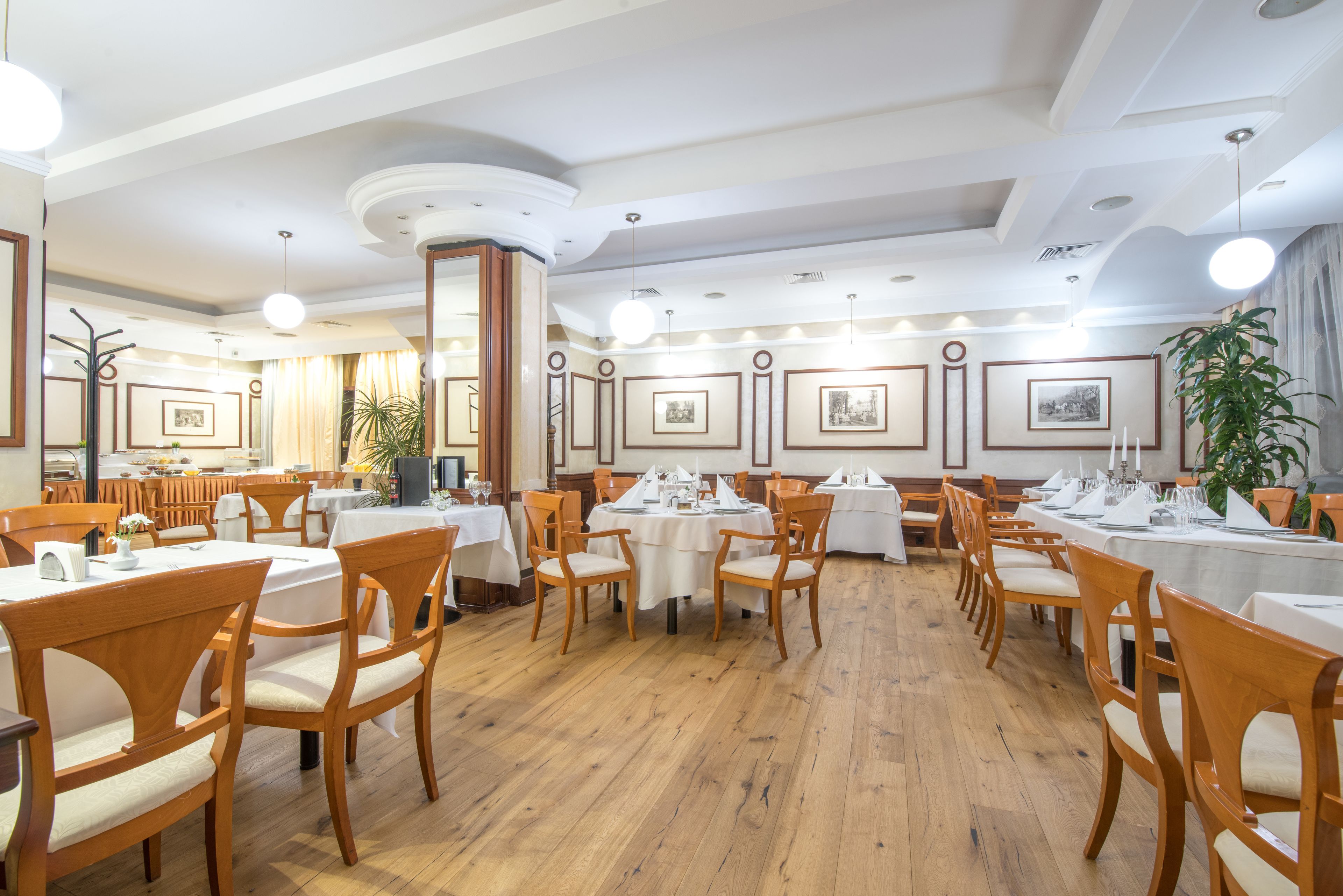 A spacious dining area with wooden chairs and tables, framed pictures on the walls, and a large potted plant.