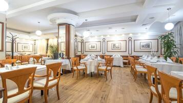 A spacious dining area with wooden chairs and tables, framed pictures on the walls, and a large potted plant.