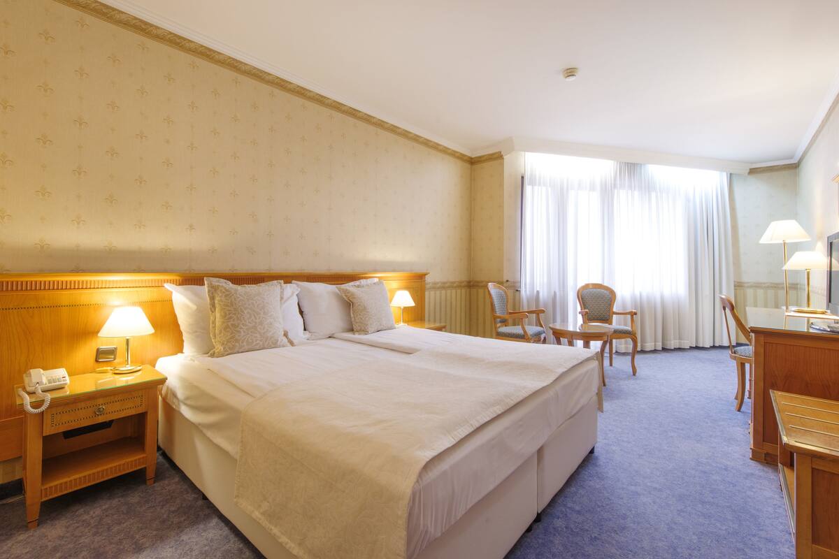 A hotel room with a large bed, a bedside table with a lamp, a desk with a chair, and a window with curtains.