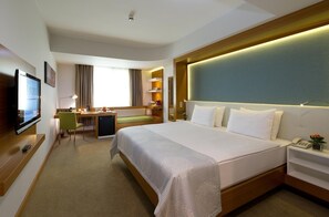 Habitación de hotel con una cama grande, un escritorio con silla, televisión y mesita de noche con teléfono y planta.