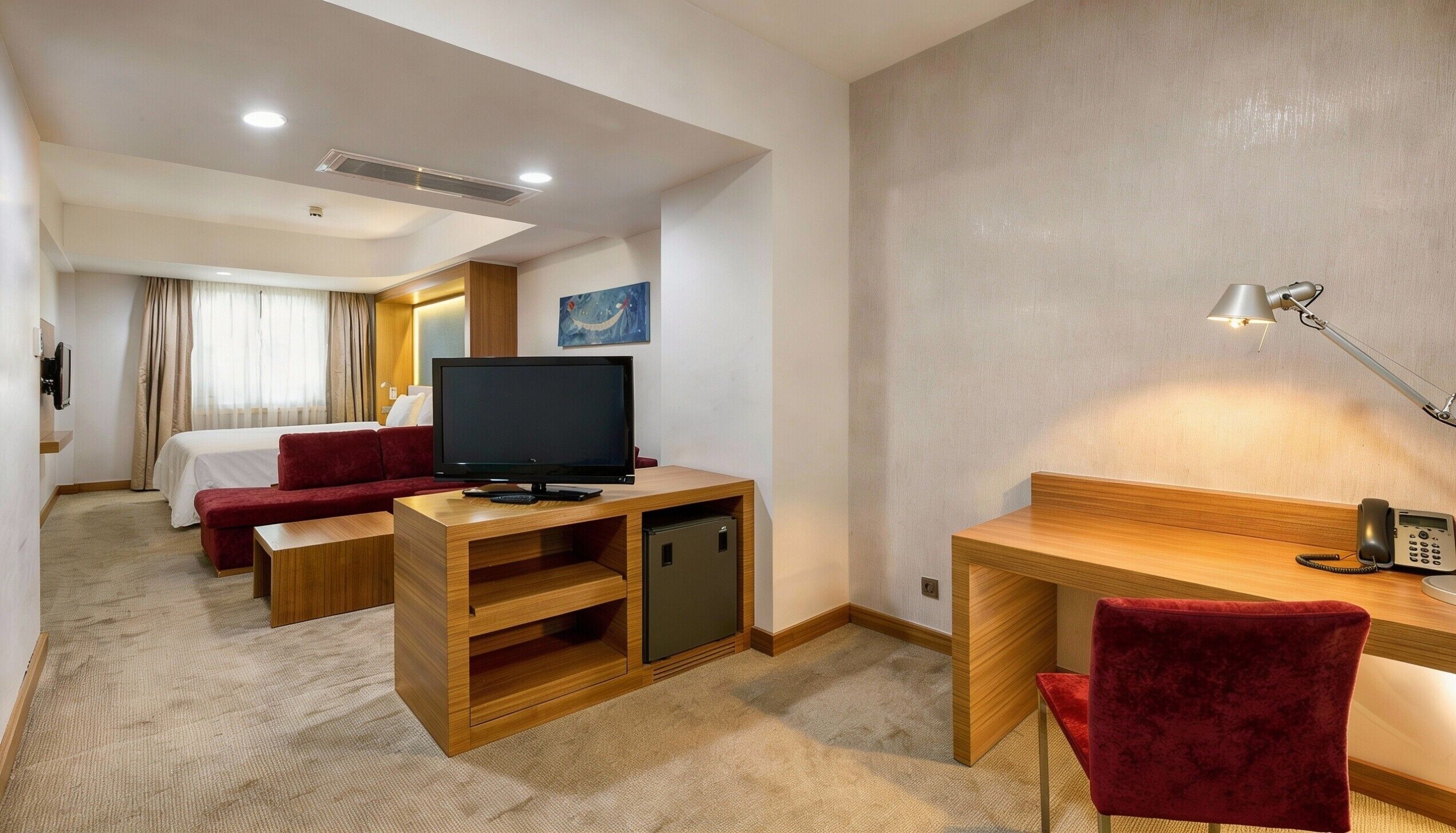 A hotel room with a bed, a TV, a desk with a lamp, a phone, and a red chair.