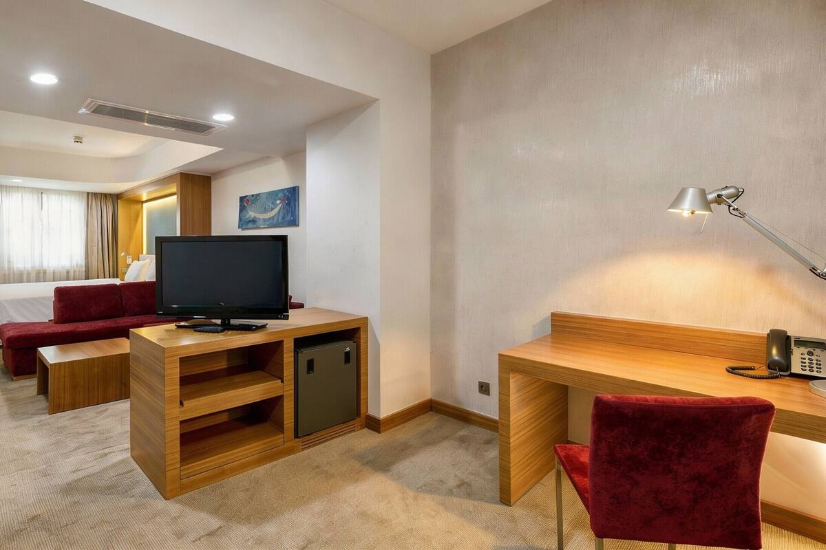 Una habitación de hotel con una cama, televisión, escritorio con lámpara, teléfono y una silla roja.