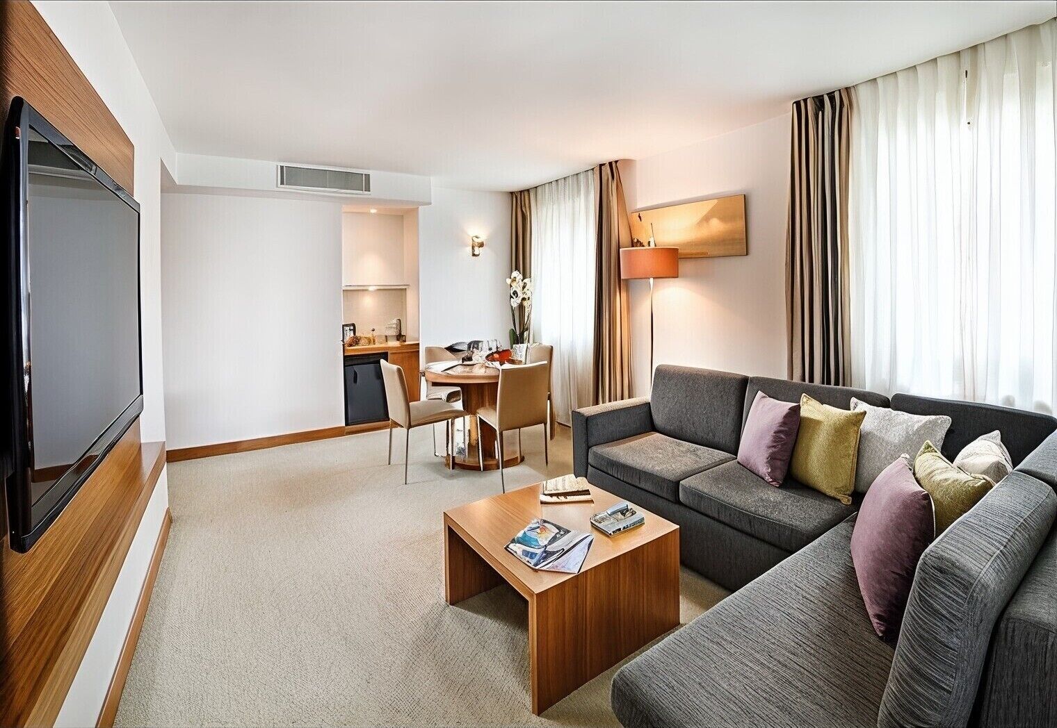 A modern hotel room with a flat-screen TV, a sofa with multiple cushions, a wooden coffee table, a dining area with a table and chairs, and a large window with curtains.