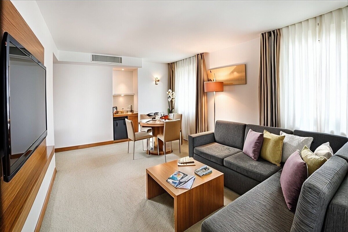A modern hotel room with a flat-screen TV, a sofa with multiple cushions, a wooden coffee table, a dining area with a table and chairs, and a large window with curtains.