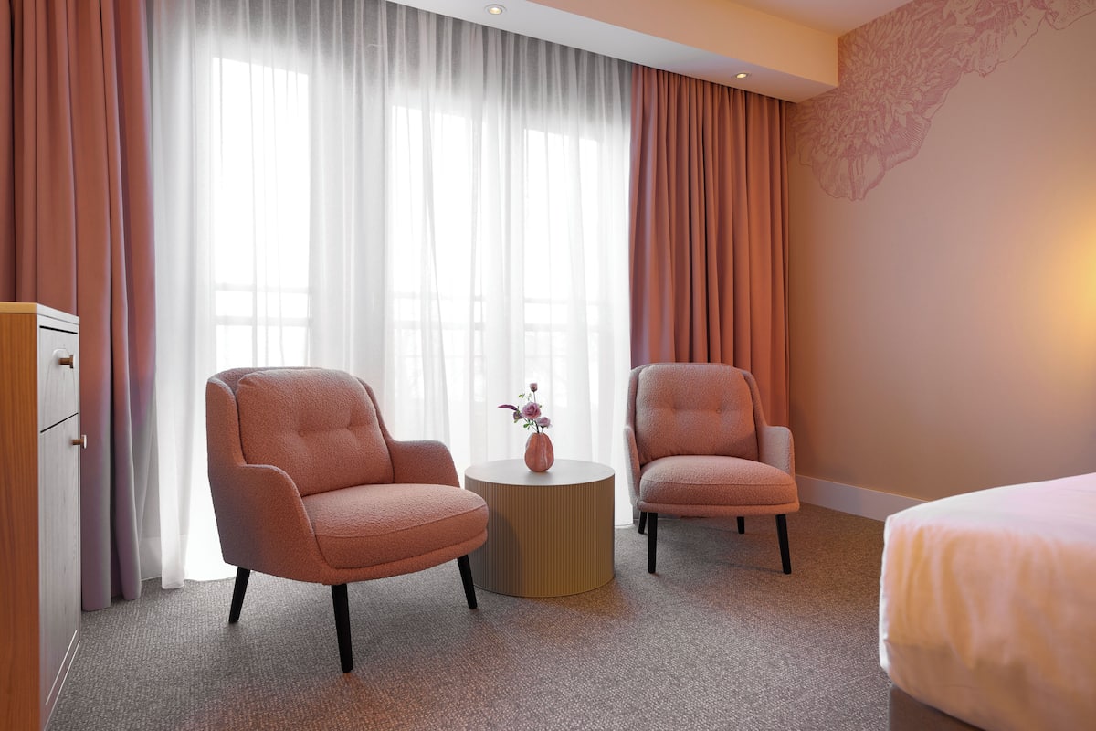 Ein Hotelzimmer mit zwei Sesseln, einem runden Beistelltisch mit einer Vase, einem Bett und einem großen Fenster mit durchsichtigen Vorhängen.