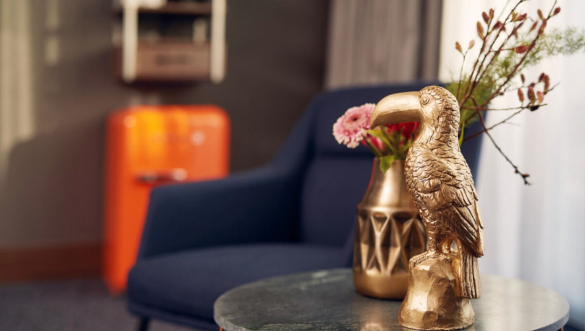 A room with a blue armchair, a small round table with a decorative golden bird and a vase with pink flowers.