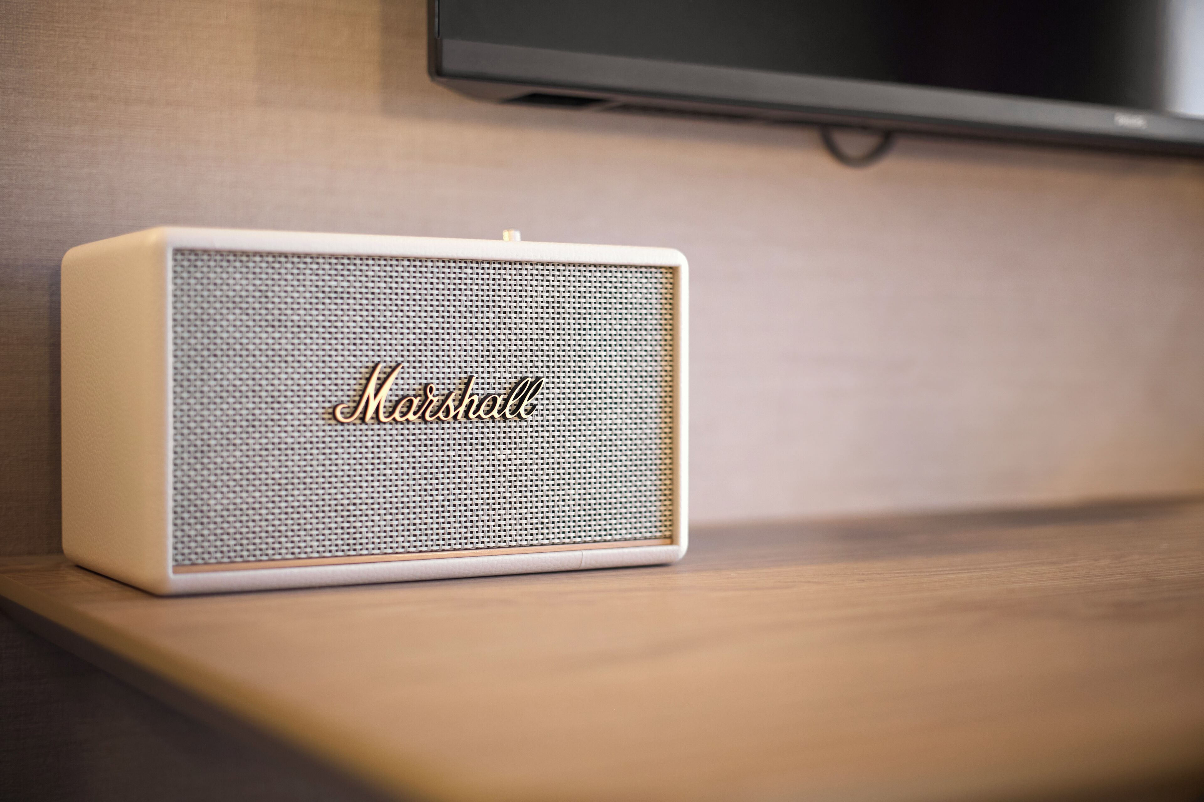 A white Marshall speaker on a wooden surface with a television mounted on the wall behind it.