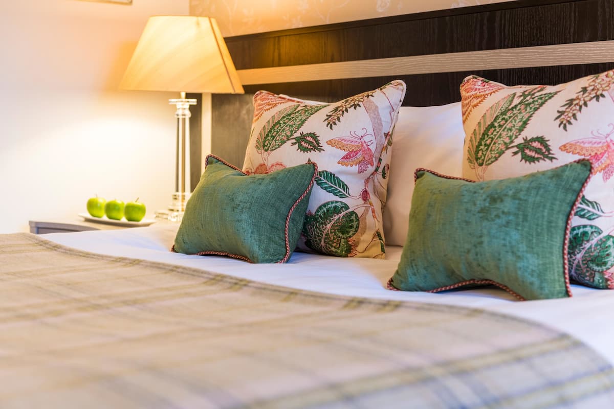 A neatly made bed with green pillows and a floral-patterned pillow, a bedside lamp, and a plate with apples.
