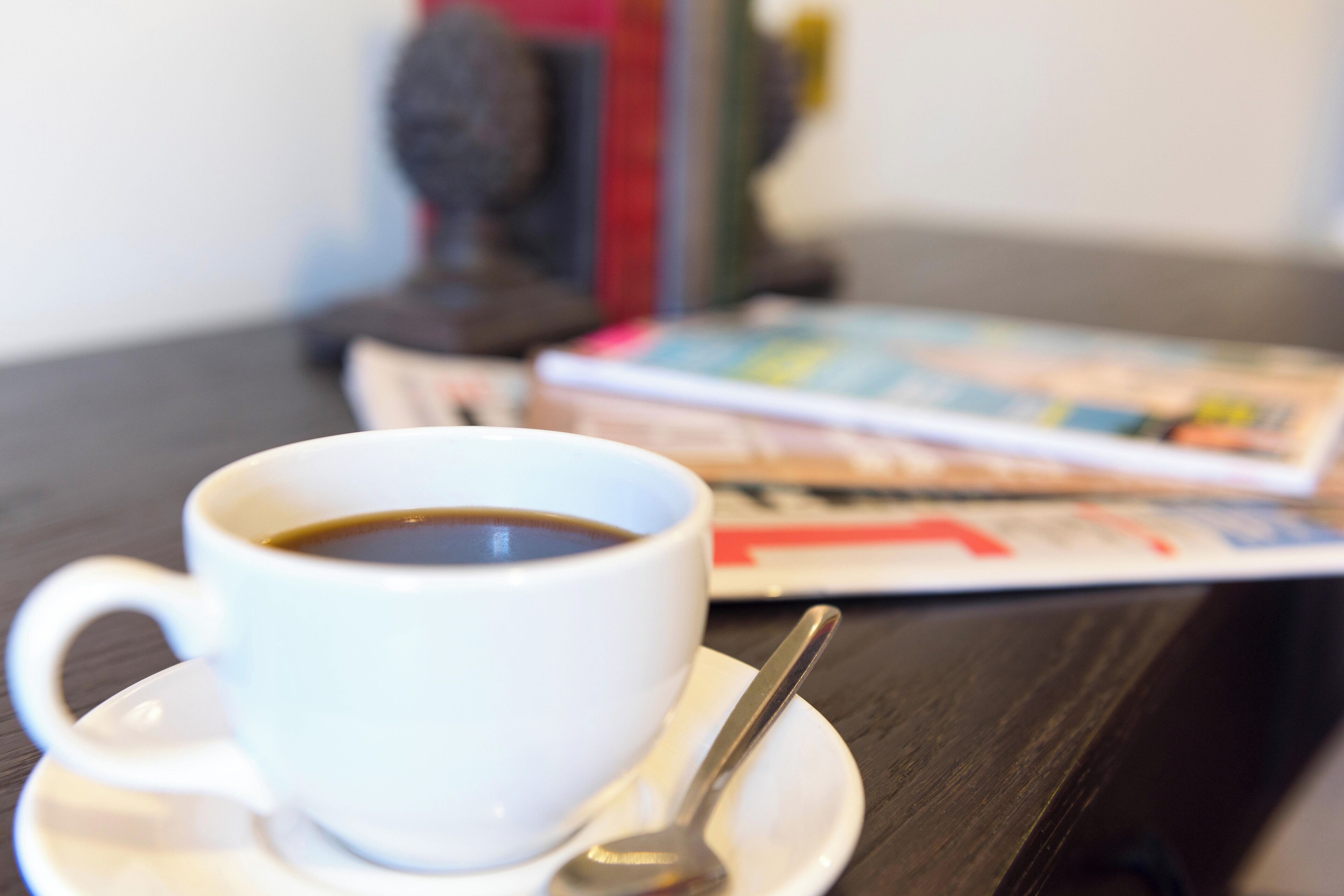 A white cup of coffee on a saucer with a spoon, a magazine, and a small statue on a dark wooden table.