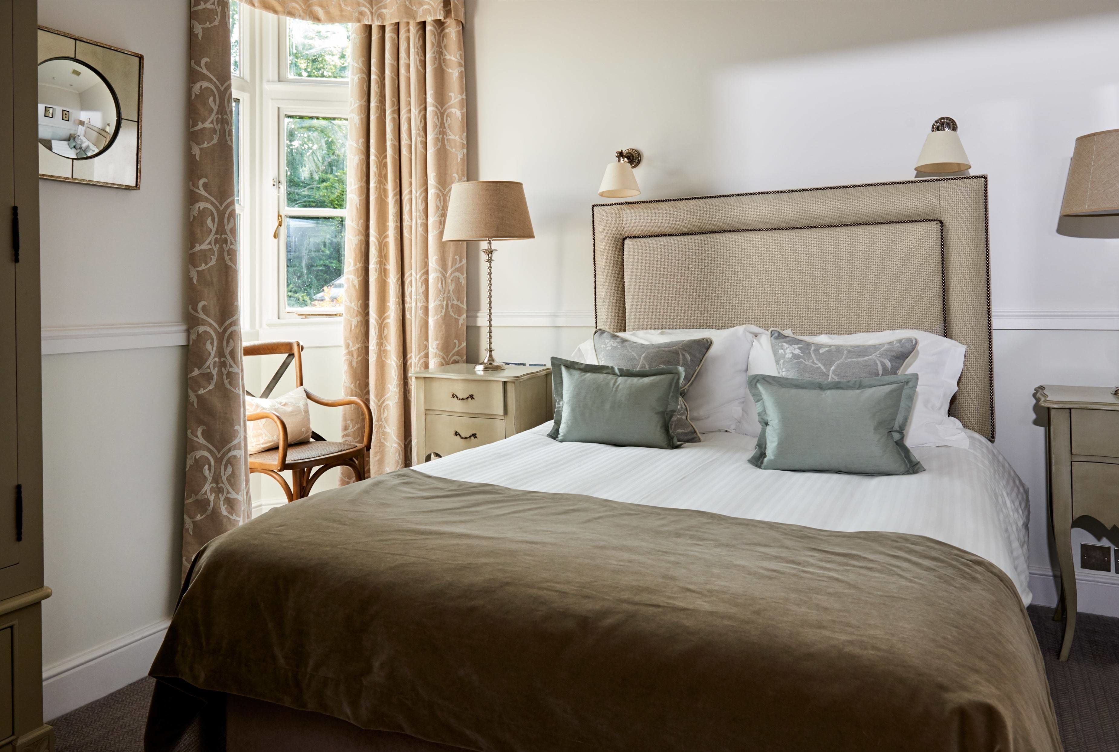 A neatly arranged bedroom with a large bed, a bedside table with a lamp, a chair, and a window with curtains.