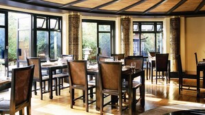 A dining area with wooden tables and chairs, large windows, and a ceiling with exposed beams. - Careys Manor & Senspa (Brockenhurst)