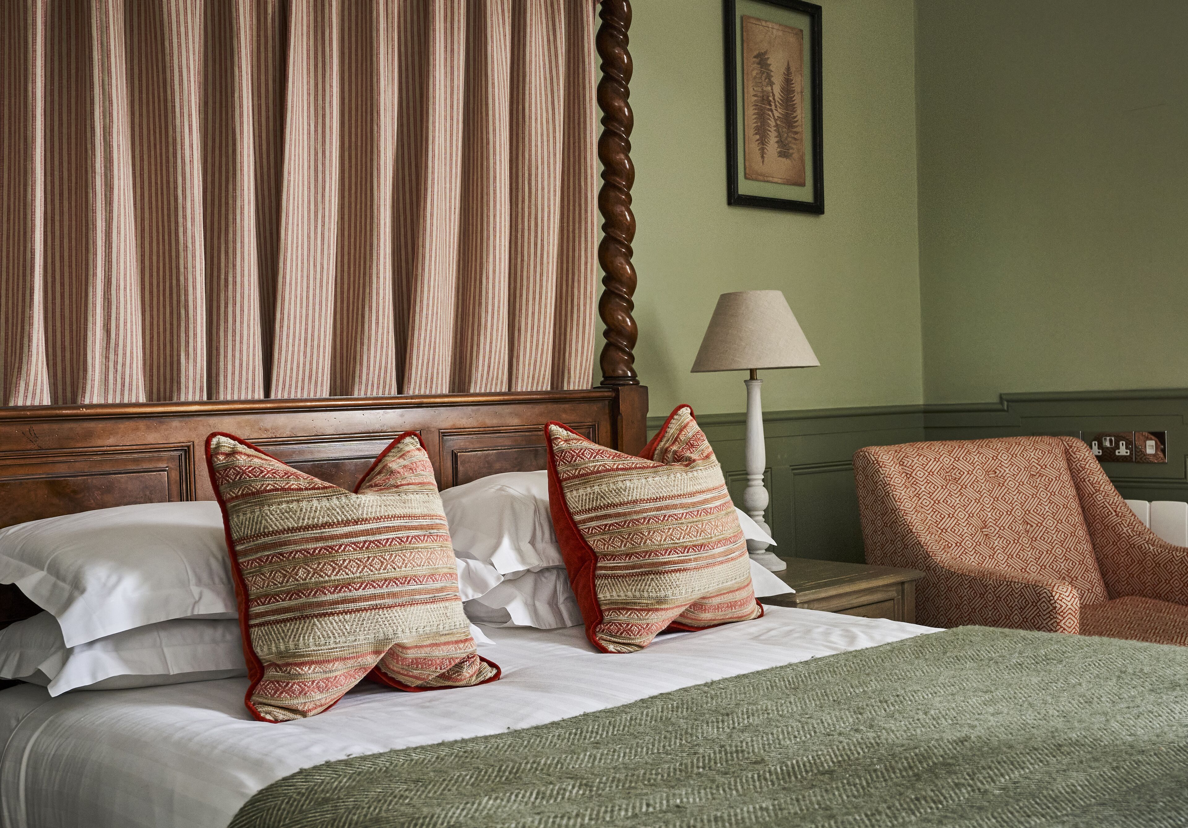 A neatly made bed with patterned pillows, a wooden headboard, a green throw blanket, a lamp, and a patterned armchair.