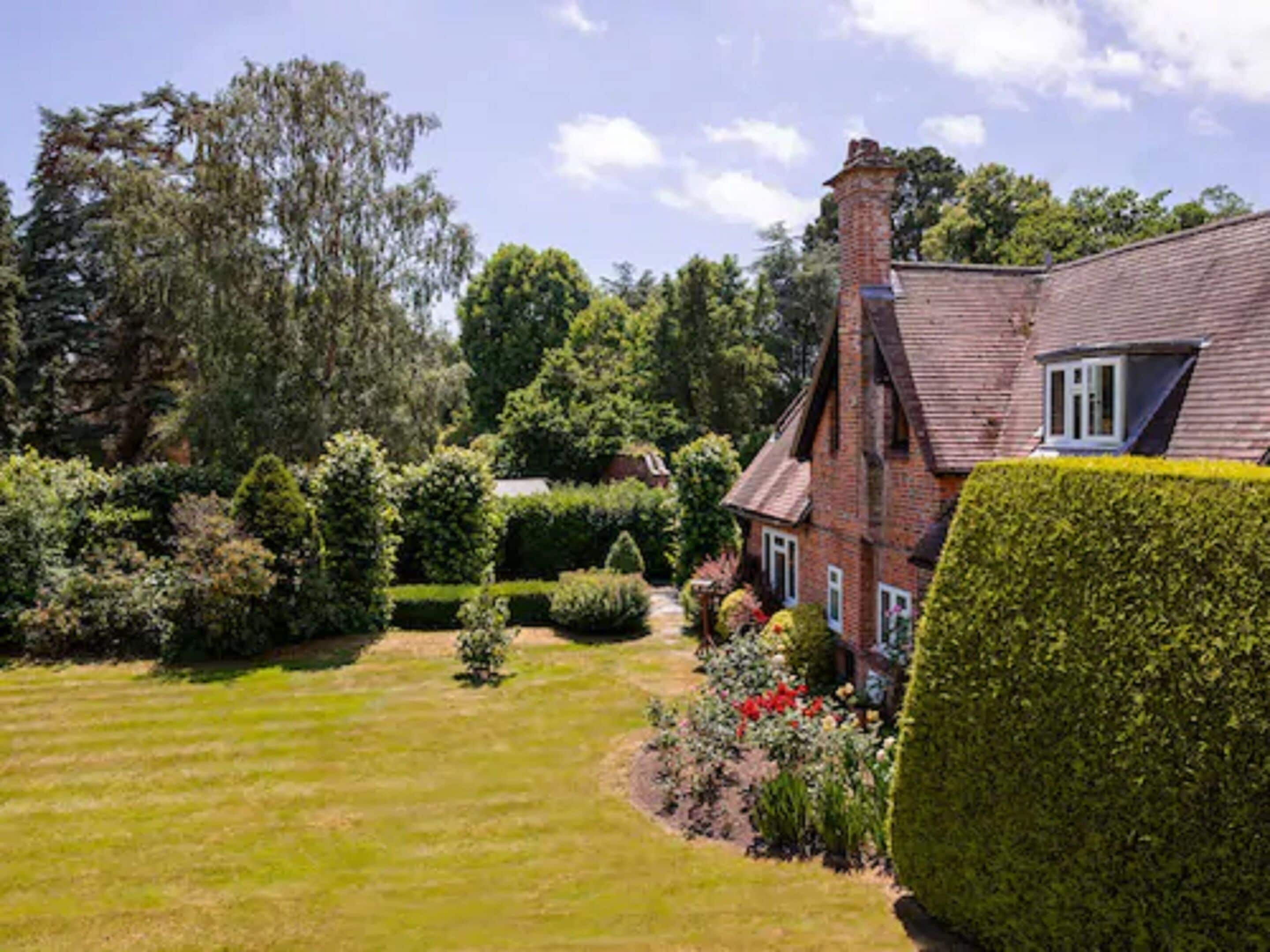 A traditional brick house with a well-maintained garden, including a lawn, hedges, and flowering plants.