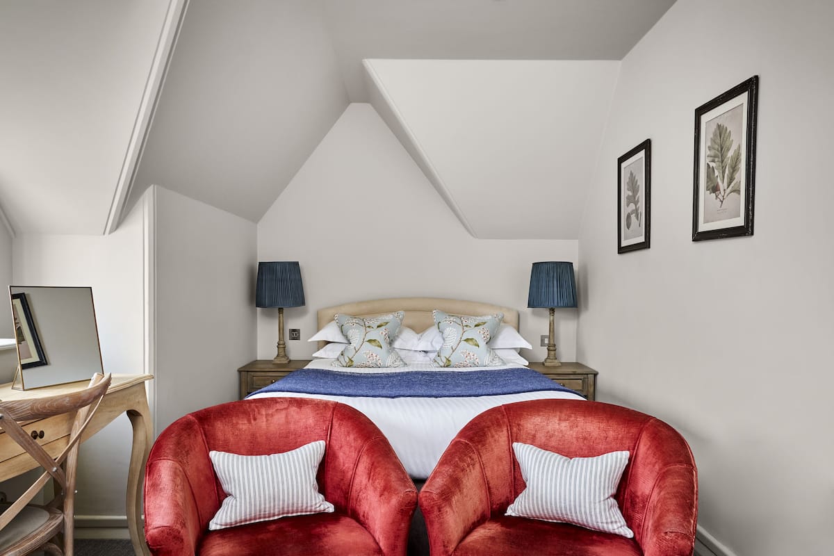 A bedroom with a large bed, two red velvet chairs, a desk, and a nightstand with lamps.