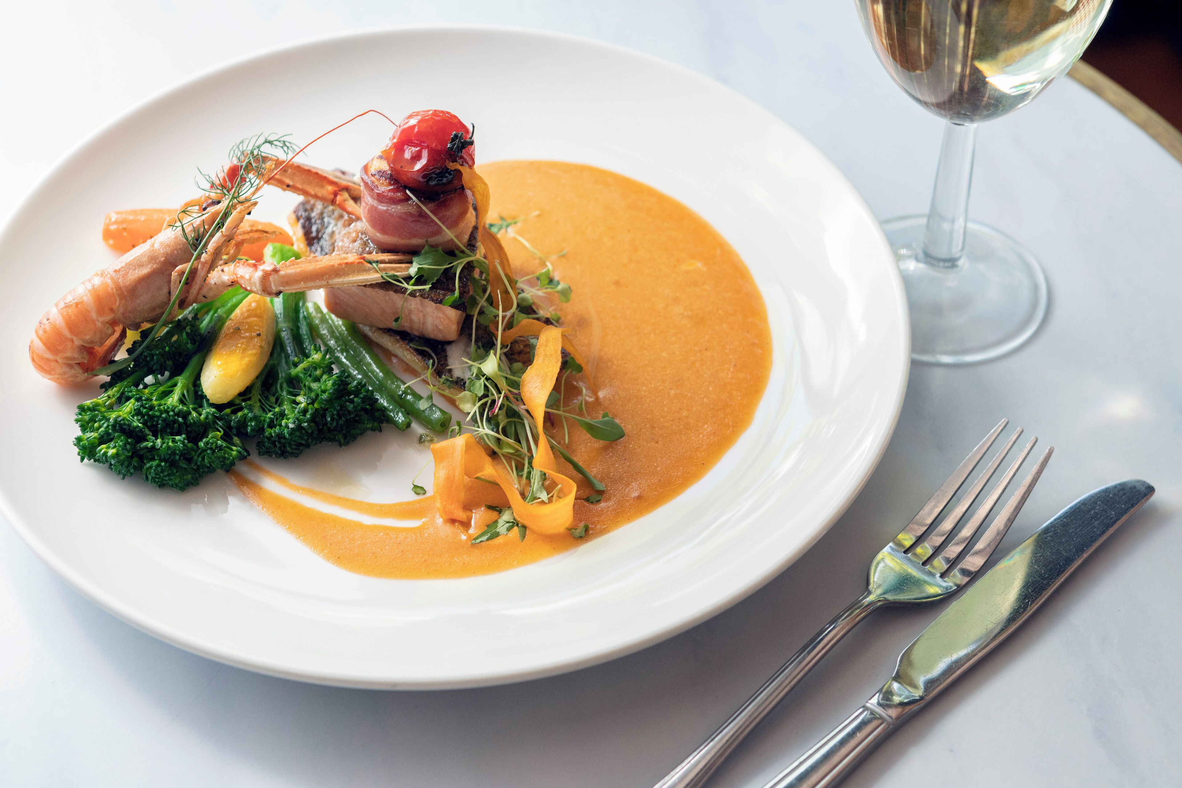 A white plate with a seafood dish, a glass of white wine, and silverware on a marble table.
