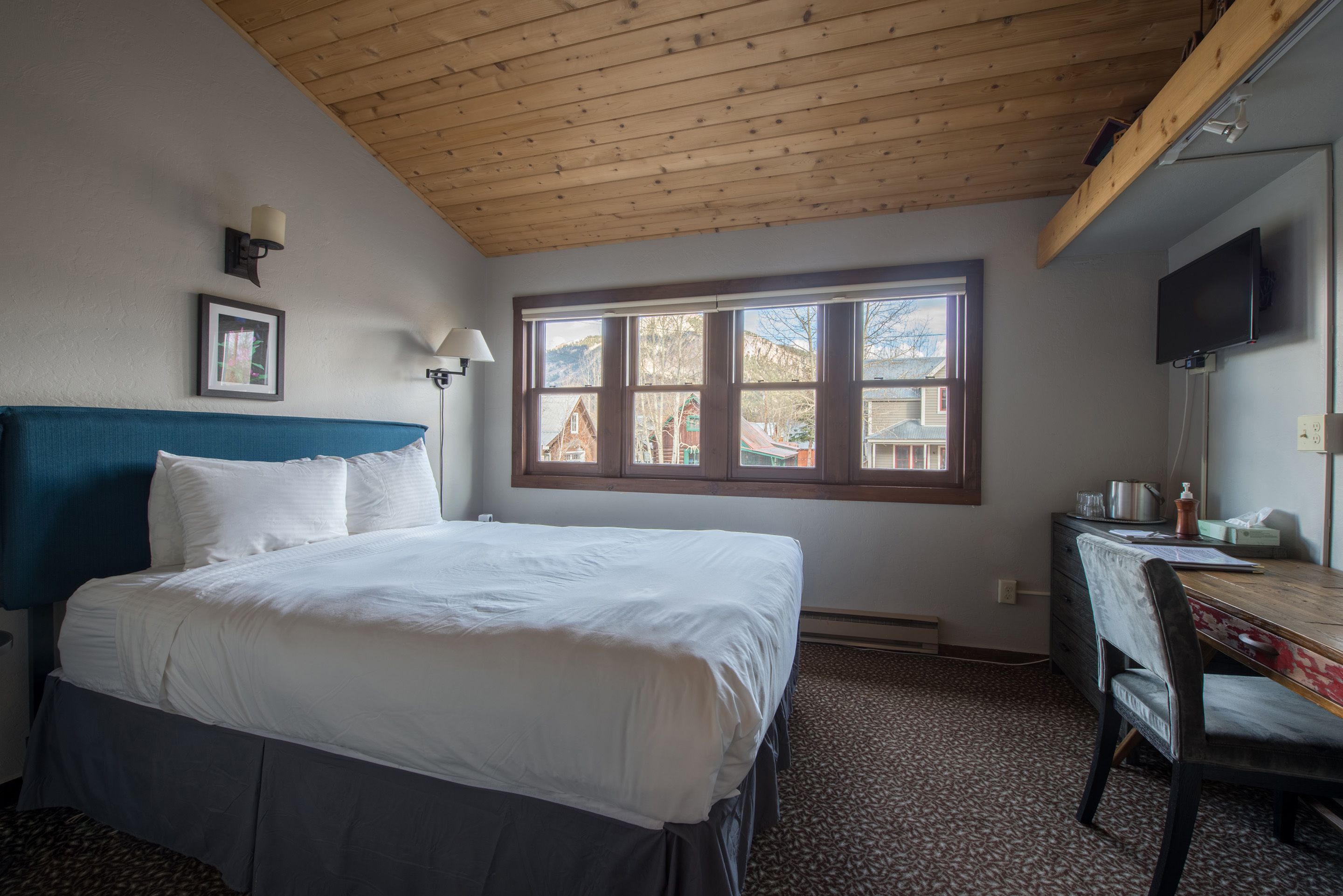 Une chambre à coucher comprenant un lit, un bureau, une chaise, un téléviseur et une fenêtre donnant sur des immeubles.