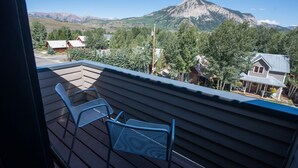 En balkon med to metalstole, udsigt over et bjerg og et boligområde med huse og træer.