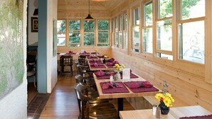 En restaurant med trævægge og store vinduer, borde dækket med lilla servietter og blomster.
