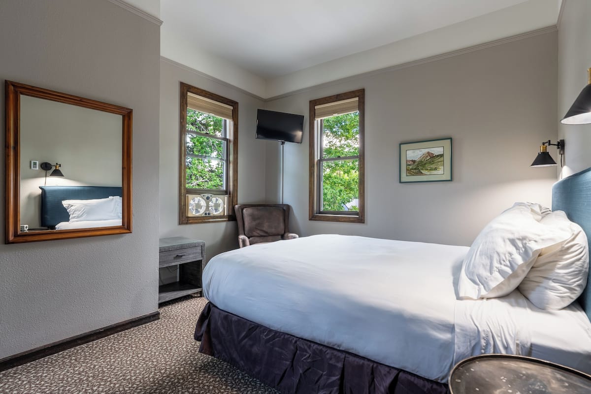Ein Hotelzimmer mit einem großen Bett, einem Schreibtisch mit Stuhl, einem Fernseher und zwei Fenstern mit Blick auf Grünflächen.