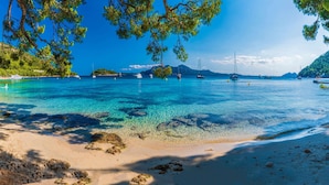 Une plage bordée d’une eau turquoise et limpide, avec des bateaux et des îles qui se dessinent à l’horizon.