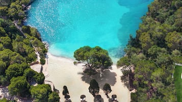 An aerial view of a beach with clear turquoise waters, surrounded by dense green trees. There are several thatched-roof umbrellas and lounge chairs arranged along the shoreline.