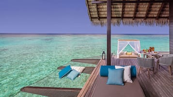 Una terraza de madera con techo de paja, un área de descanso acolchada con almohadones y una mesa lista para comer, con vistas a un mar turquesa cristalino.